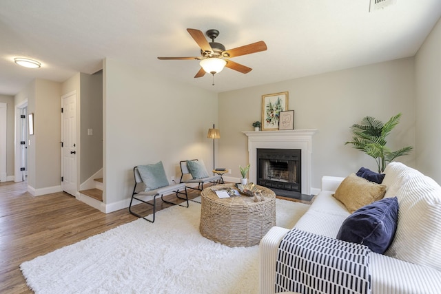 living area with a fireplace with flush hearth, a ceiling fan, wood finished floors, baseboards, and stairs