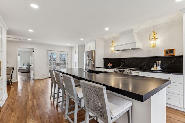 kitchen with a center island with sink, custom range hood, a sink, dark countertops, and light wood finished floors