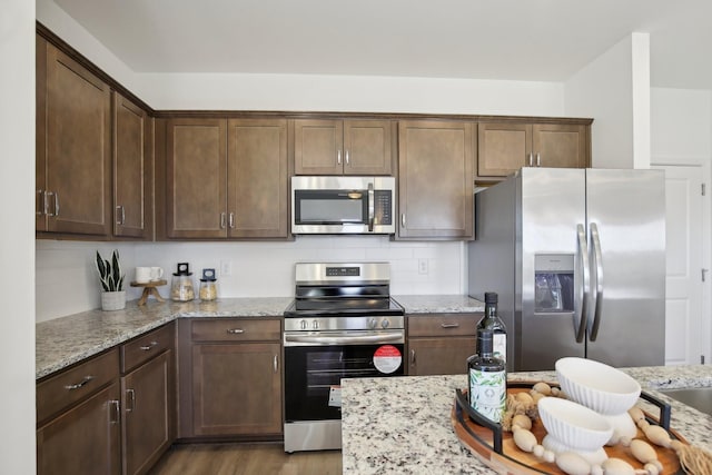 kitchen with stainless steel appliances, tasteful backsplash, dark brown cabinetry, wood finished floors, and light stone countertops