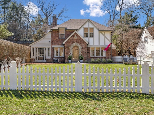 tudor-style house featuring stucco siding, a chimney, a front lawn, a fenced front yard, and brick siding