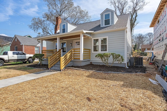 new england style home featuring a porch, roof with shingles, central AC, and a chimney