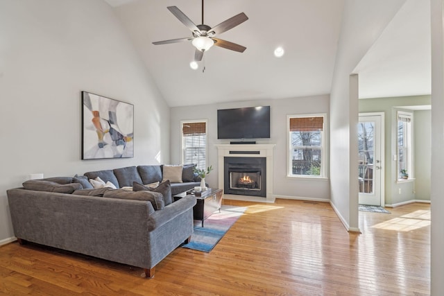 living area with light wood-type flooring, a fireplace with flush hearth, high vaulted ceiling, baseboards, and ceiling fan