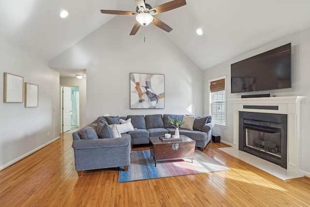 living room featuring baseboards, high vaulted ceiling, a fireplace with flush hearth, ceiling fan, and light wood-type flooring