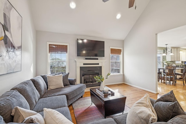 living area featuring baseboards, light wood-type flooring, a glass covered fireplace, high vaulted ceiling, and a ceiling fan