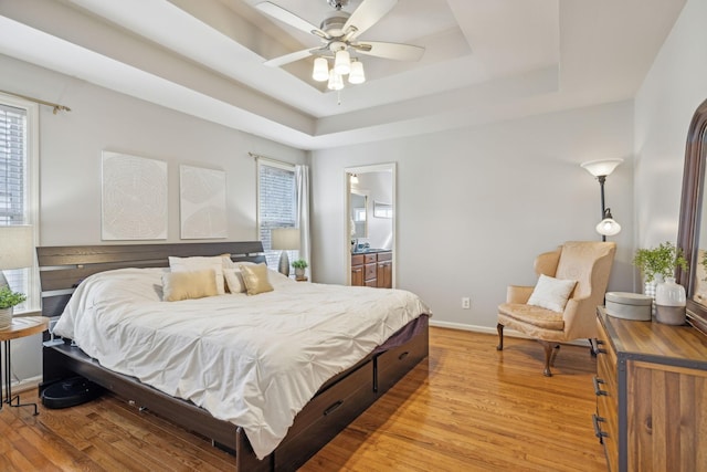 bedroom featuring a tray ceiling, baseboards, light wood-style floors, and ensuite bath