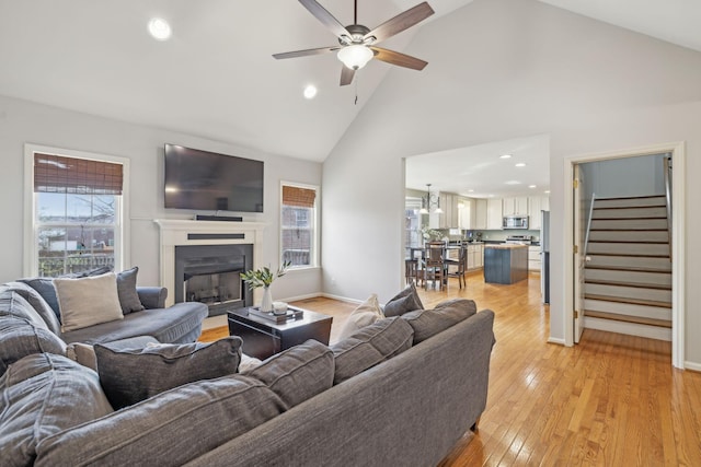 living area featuring light wood-type flooring, a fireplace, baseboards, ceiling fan, and stairs