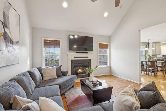 living area with ceiling fan, baseboards, light wood-style floors, a glass covered fireplace, and high vaulted ceiling