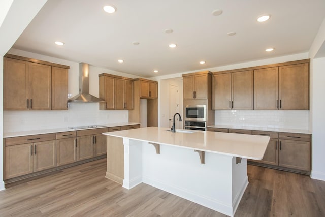 kitchen with brown cabinets, a sink, wood finished floors, appliances with stainless steel finishes, and wall chimney exhaust hood