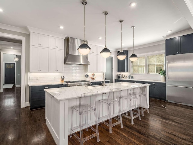 kitchen featuring dark wood-style floors, stainless steel appliances, wall chimney exhaust hood, decorative backsplash, and a large island with sink