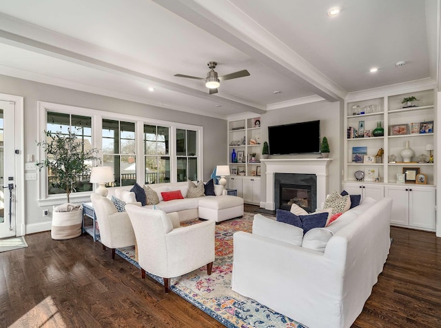 living room with dark wood-type flooring, built in features, beam ceiling, a glass covered fireplace, and a ceiling fan