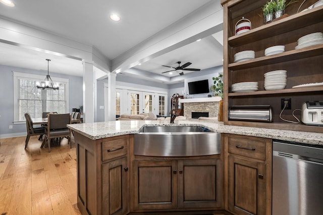 kitchen featuring light wood finished floors, a stone fireplace, ceiling fan with notable chandelier, stainless steel dishwasher, and a sink