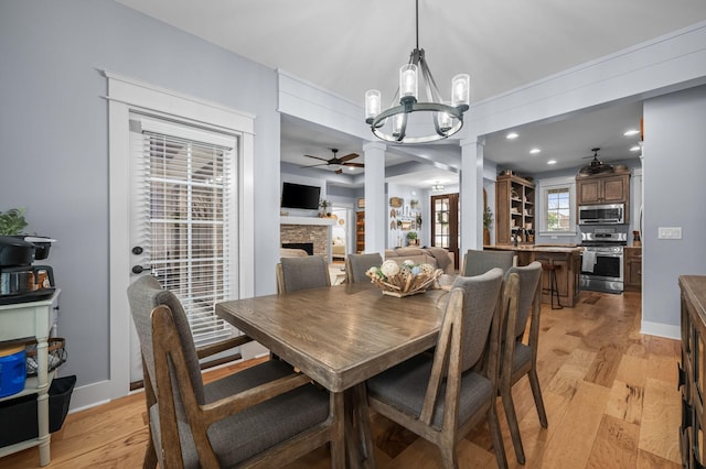 dining space featuring baseboards, ornate columns, a fireplace, ceiling fan with notable chandelier, and light wood-type flooring