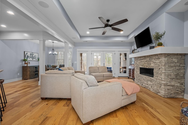 living area with a stone fireplace, light wood-style flooring, decorative columns, and ceiling fan with notable chandelier