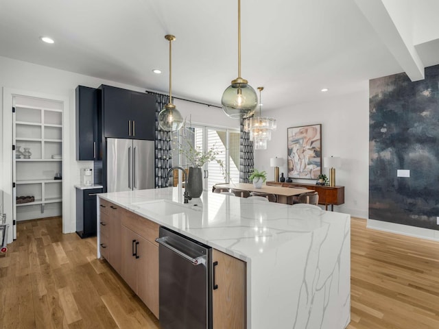 kitchen with appliances with stainless steel finishes, light stone countertops, a sink, light wood-style floors, and an island with sink