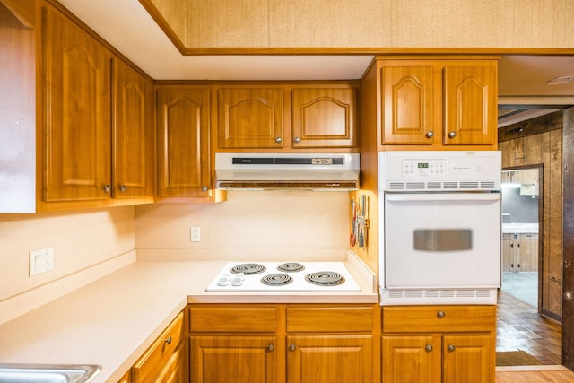 kitchen with under cabinet range hood, brown cabinetry, white appliances, and light countertops