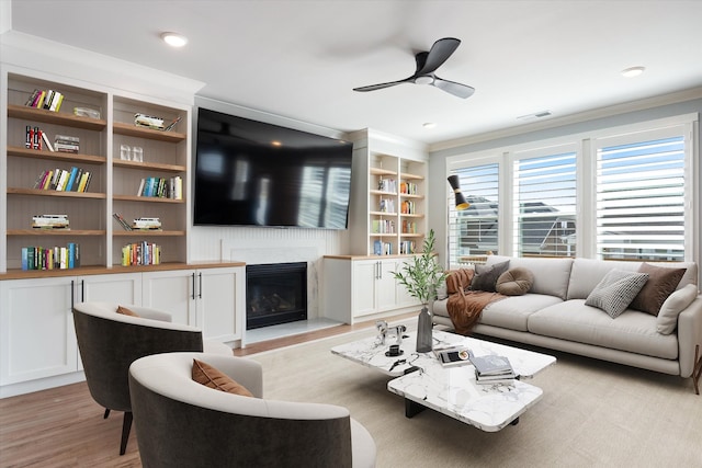 living area featuring visible vents, built in features, a glass covered fireplace, light wood-style floors, and ceiling fan