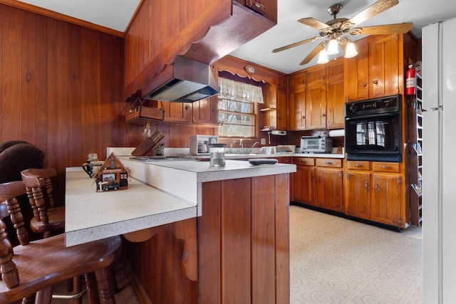 kitchen featuring black oven, wood walls, light countertops, brown cabinets, and a ceiling fan