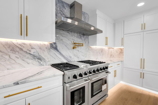 kitchen with light stone countertops, range with two ovens, decorative backsplash, white cabinets, and wall chimney range hood