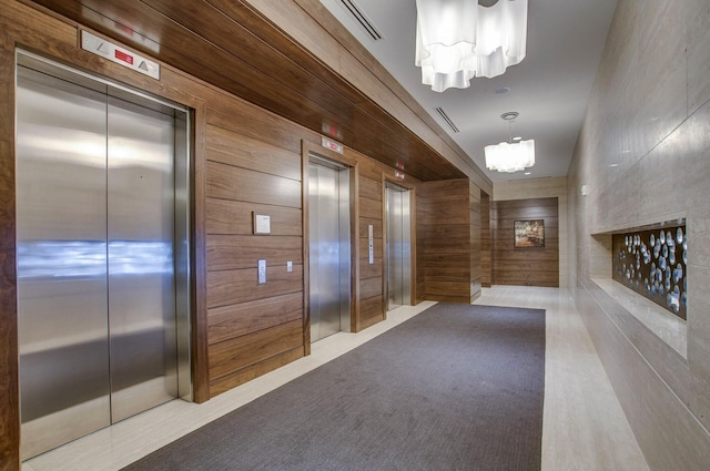 hallway with elevator, a notable chandelier, wood walls, and visible vents