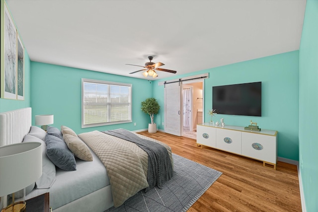 bedroom with wood finished floors, baseboards, ceiling fan, a barn door, and connected bathroom