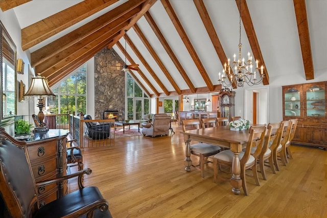 dining room with light wood-type flooring, beam ceiling, high vaulted ceiling, a stone fireplace, and an inviting chandelier