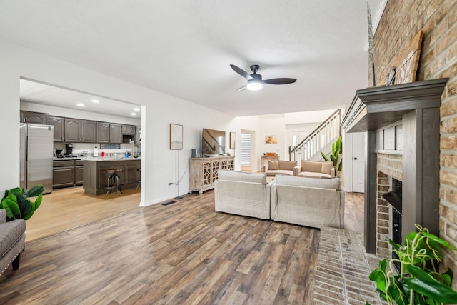 living area with a brick fireplace, baseboards, stairs, wood finished floors, and a ceiling fan