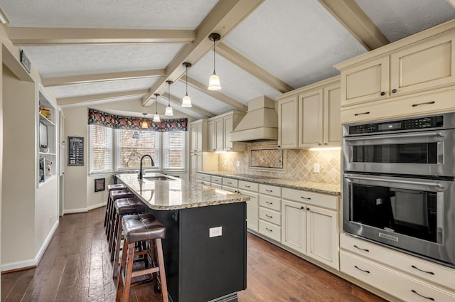 kitchen with stainless steel double oven, custom exhaust hood, a sink, cream cabinets, and tasteful backsplash