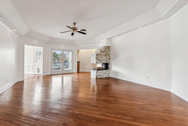 unfurnished living room featuring baseboards, ceiling fan, hardwood / wood-style floors, a stone fireplace, and a raised ceiling