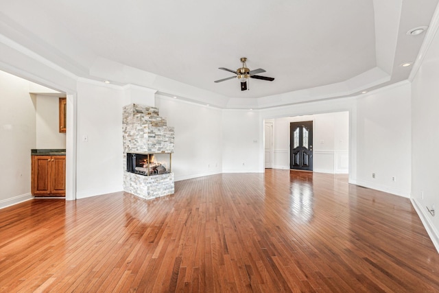 unfurnished living room with a ceiling fan, baseboards, a tray ceiling, a stone fireplace, and light wood-style floors