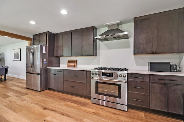 kitchen with dark brown cabinets, wall chimney range hood, light countertops, light wood-style floors, and stainless steel appliances