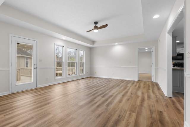 unfurnished living room with a ceiling fan, a tray ceiling, baseboards, and light wood finished floors