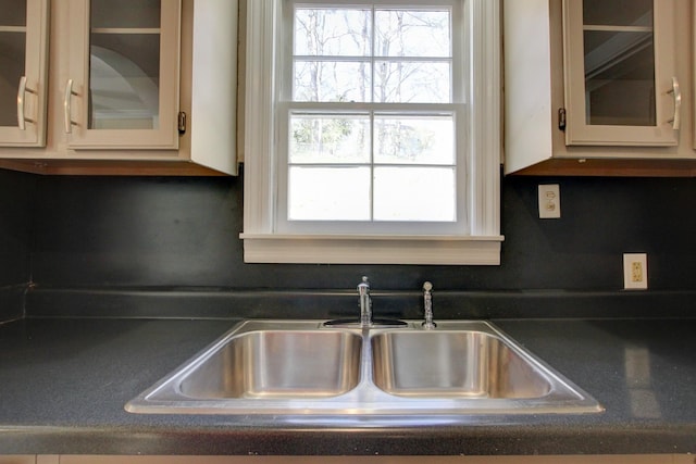 kitchen with a sink, glass insert cabinets, dark countertops, and cream cabinetry