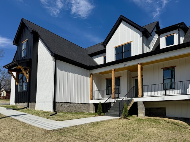 view of side of home featuring brick siding, board and batten siding, a porch, roof with shingles, and a lawn