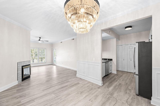 unfurnished living room featuring a tiled fireplace, light wood-type flooring, ornamental molding, ceiling fan with notable chandelier, and wainscoting