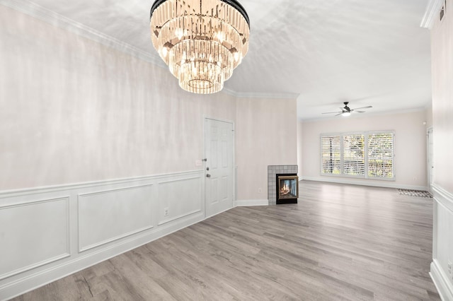 unfurnished living room with wood finished floors, a tile fireplace, crown molding, a decorative wall, and ceiling fan with notable chandelier