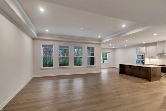 unfurnished living room featuring a tray ceiling, wood finished floors, baseboards, a sink, and crown molding