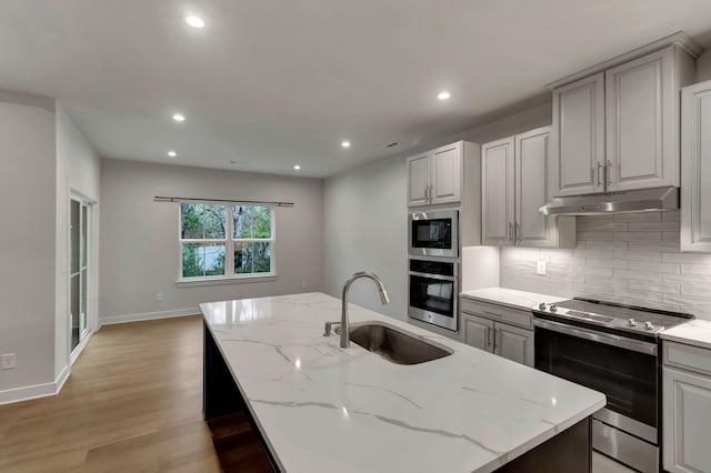 kitchen featuring appliances with stainless steel finishes, a sink, under cabinet range hood, light wood-style flooring, and backsplash