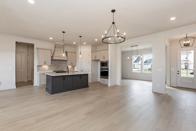kitchen with tasteful backsplash, a notable chandelier, appliances with stainless steel finishes, premium range hood, and a kitchen island with sink