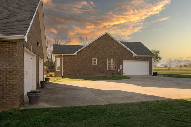 property exterior at dusk with an attached garage, concrete driveway, brick siding, a shingled roof, and a yard