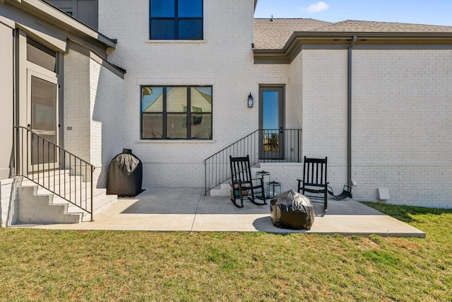 view of exterior entry featuring a lawn, brick siding, roof with shingles, and a patio area