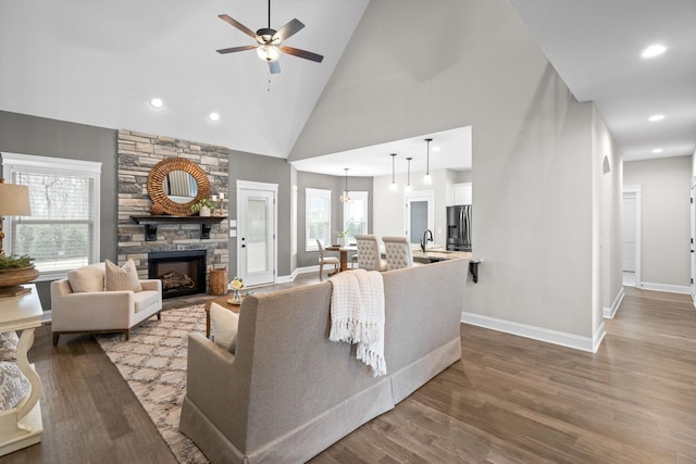 living room featuring high vaulted ceiling, baseboards, ceiling fan, a stone fireplace, and dark wood-type flooring