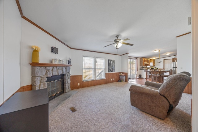 carpeted living area with wood walls, crown molding, a wainscoted wall, a ceiling fan, and a stone fireplace
