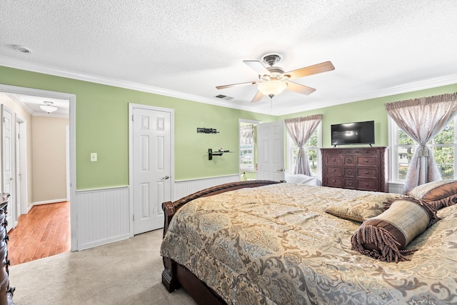 bedroom featuring a wainscoted wall, a textured ceiling, light colored carpet, visible vents, and ornamental molding