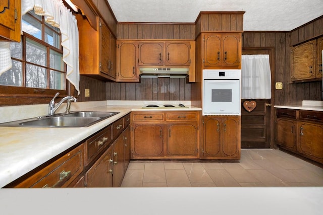 kitchen with light countertops, a sink, wood walls, under cabinet range hood, and white appliances