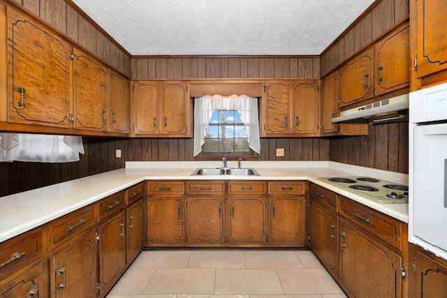 kitchen with a sink, a textured ceiling, brown cabinets, under cabinet range hood, and white appliances