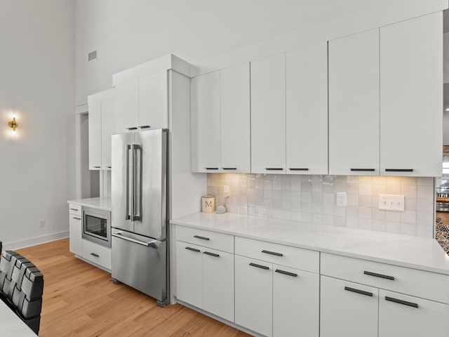 kitchen with stainless steel appliances, light wood-type flooring, a towering ceiling, backsplash, and light countertops