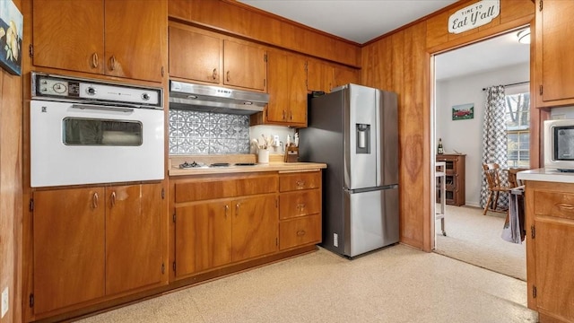 kitchen featuring crown molding, light countertops, brown cabinetry, under cabinet range hood, and white appliances