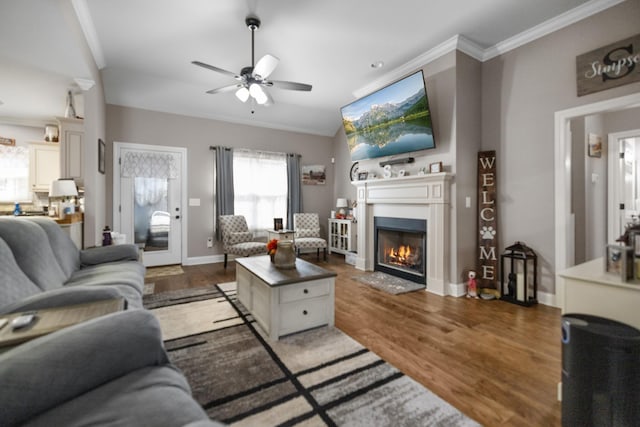 living room with crown molding, baseboards, a lit fireplace, ceiling fan, and wood finished floors