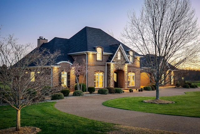 french country style house with a chimney, brick siding, curved driveway, and a front lawn
