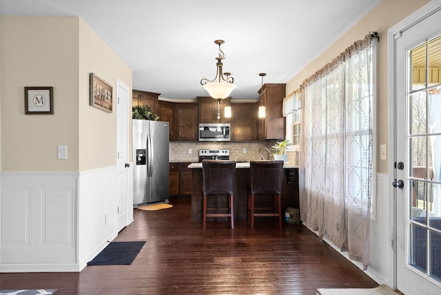 kitchen with a kitchen bar, a peninsula, wainscoting, stainless steel appliances, and dark wood-style flooring